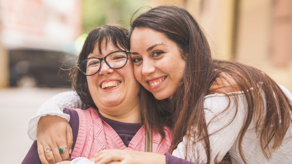 A female caregiver reaches over to hug a woman who is wearing glasses and smiling.