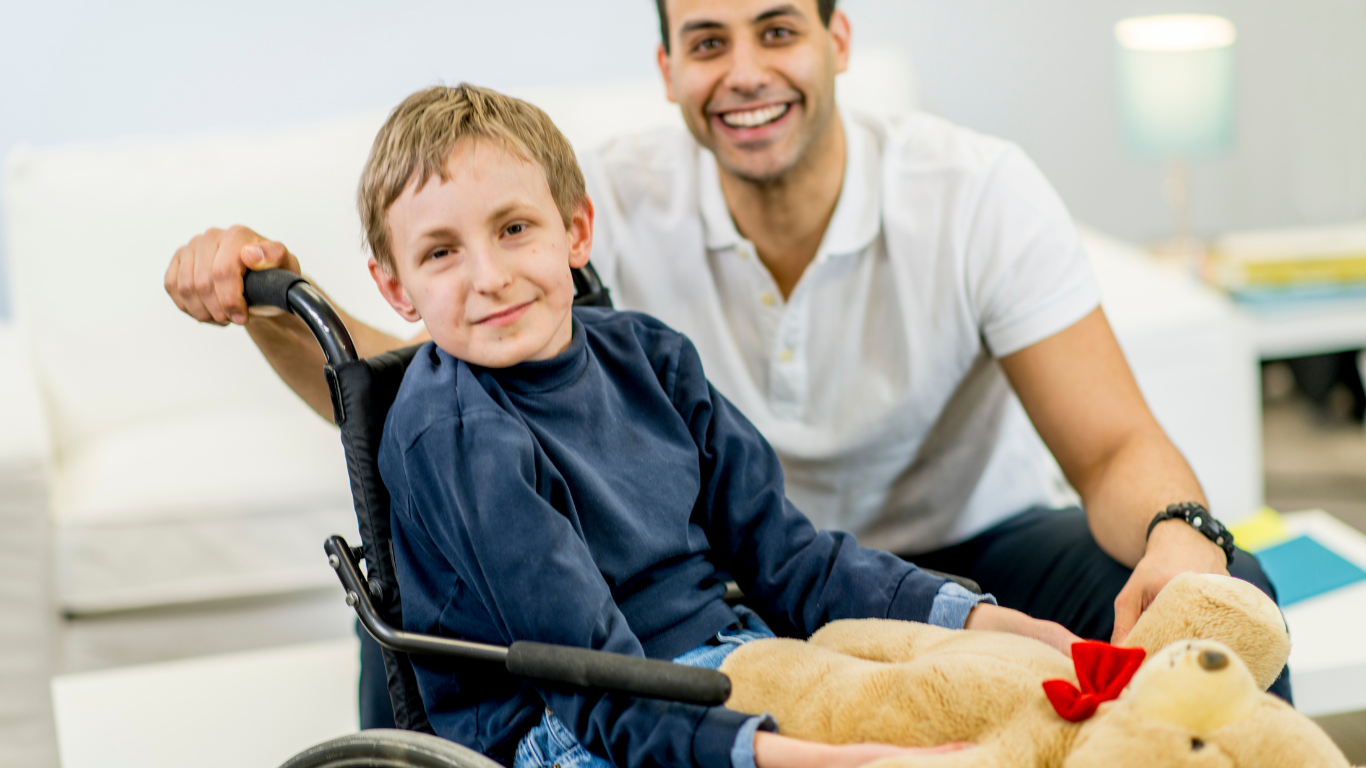 A male caregiver poses with a child in a wheelchair who holds a teddy bear with a red bow. They are both smiling.