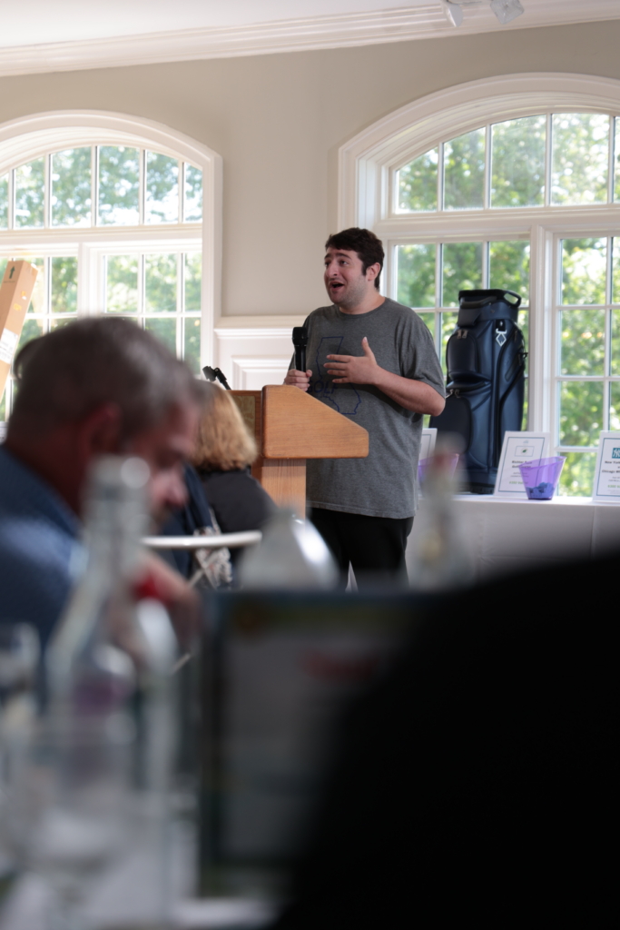 Steven stands at the podium with microphone in hand as he casually speaks to the audience about his journey to securing employment. He stands in front of large windows and speaks to guests who are seated at elegant white clothed tables.