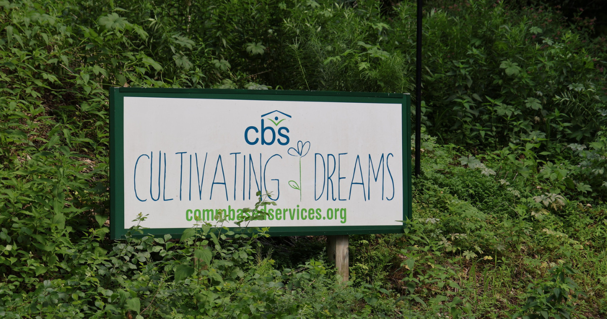The sign at Cultivating Dreams Farm with lettering on a white background. The sign is nestled into the lush greenery that grows wild around the farm.