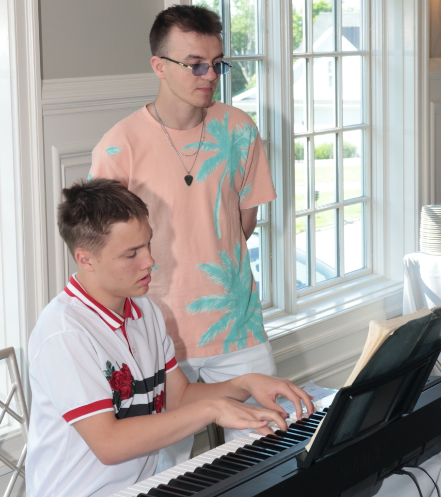 A person supported by Community Based Services performs lively and complex compositions on the piano to the delight of the audience. His teacher looks on.