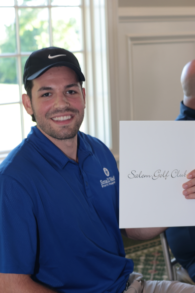 A man who had the winning bid for a Salem Golf Club gift certificated holds up his prize and smiles. He is wearing a blue ball cap and a blue collared shirt.