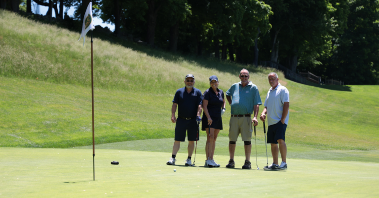 A golf foursome of three men and a woman stand on the green of Salem Golf Club smiling for th camera. A pin hole flag waves in the breeze in the foreground.