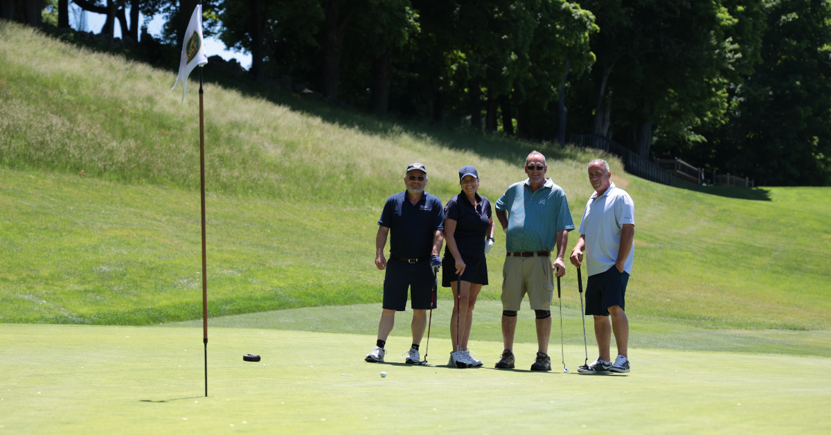 A golf foursome of three men and a woman stand on the green of Salem Golf Club smiling for th camera. A pin hole flag waves in the breeze in the foreground.
