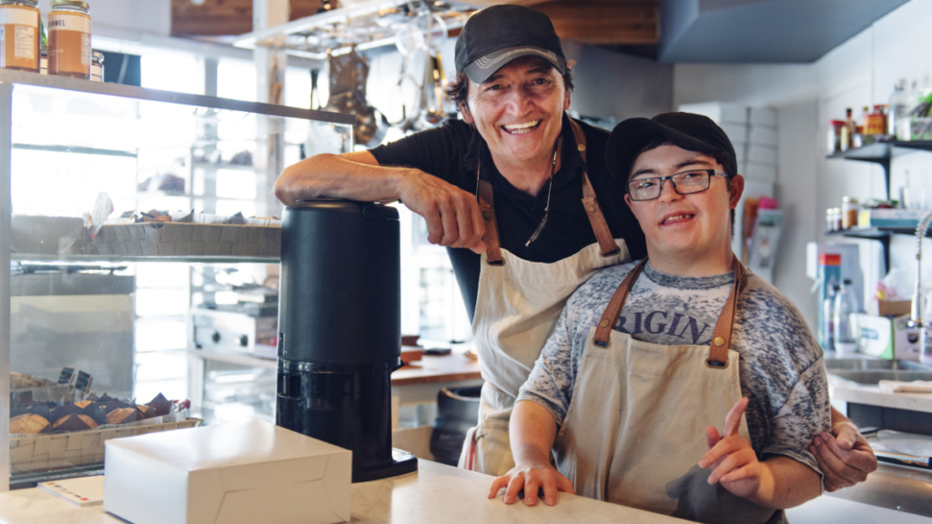 A person job shadows in a coffee shop. He wears an apron and stands next to another worker who also wears an apron. They both wear ball caps and stand at the counter by a coffee machine.
