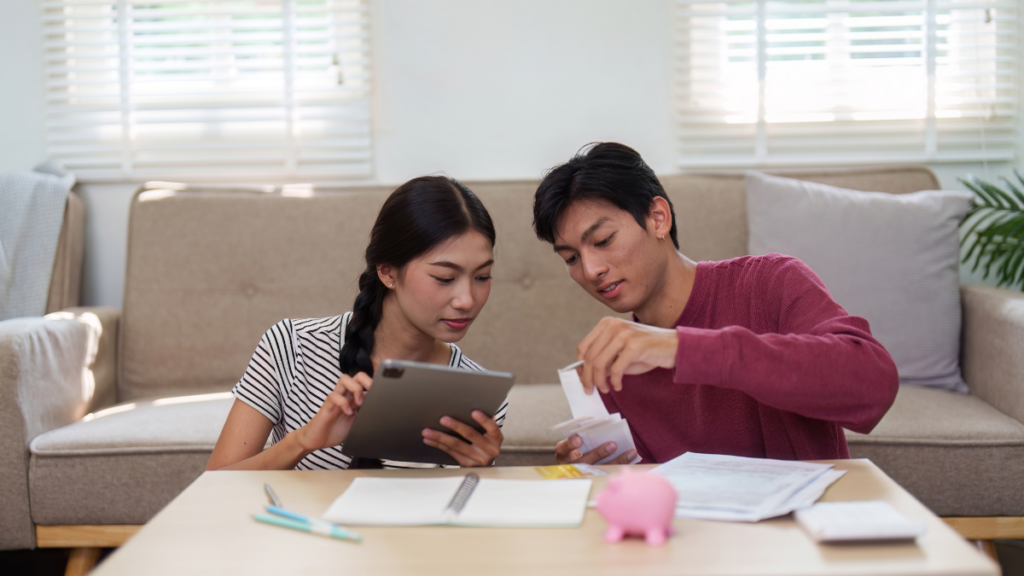Two parents sit in their living room and review documents on a tablet screen. Paper , pens and other planning supplies are spread out on the coffee table.