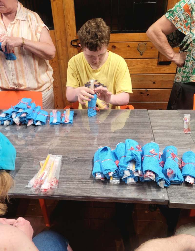 A man in a yellow shirt sits at a table in the Cultivating Dreams barn as he packs a toothbrush into a rolled up washcloth with a red ribbon tied around it.