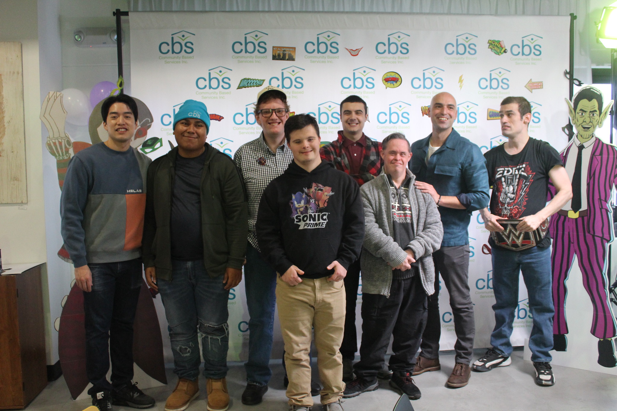 The CBS creators of the Zero to Hero comic book pose for a group photo in front of a CBS backdrop. Life-sized characters from the comic book stand on either side of the group.