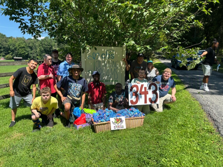 The CBS Group Day Hab poses in front of the Cultivating Dreams sign at the farm. In front of them are a large box of bags they packed for donations and one young man holds a sign with the number 343 painted on it.
