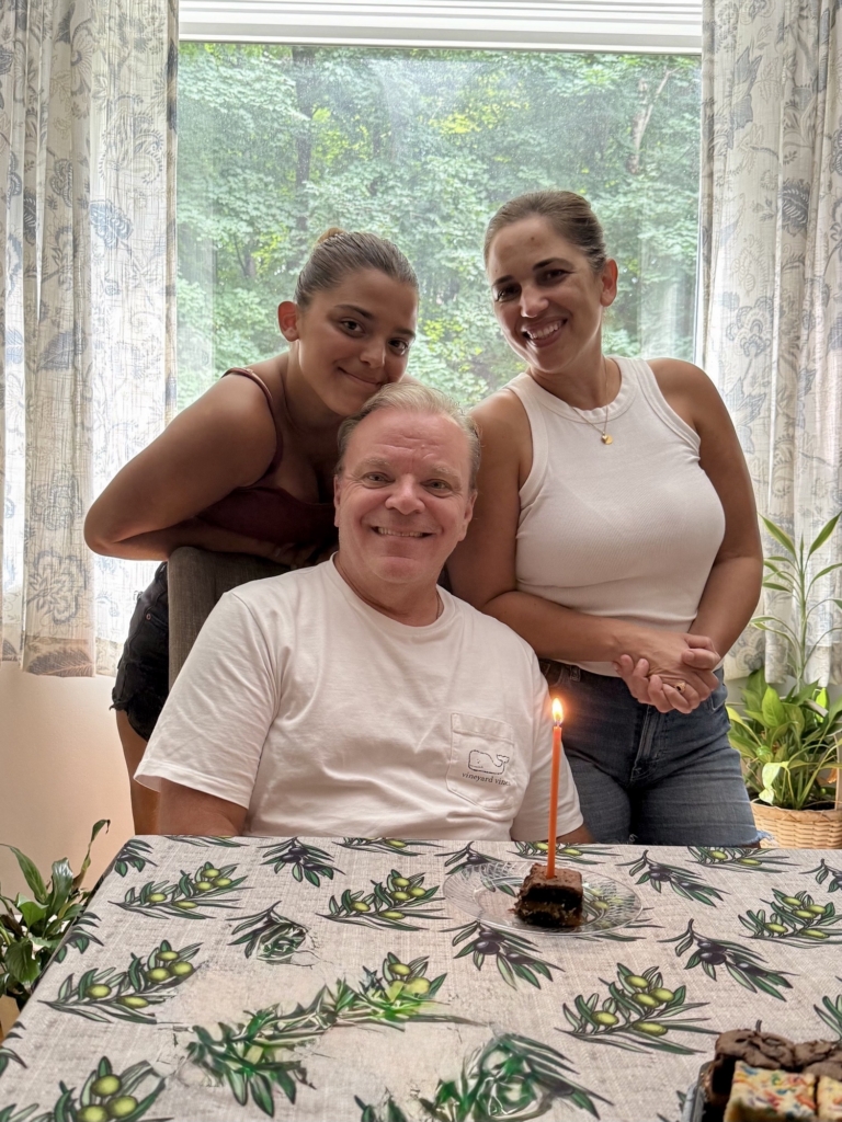 Magda Piacentino stands with another woman behind a seated man at a kitchen table. There is a cupcake and candle in front of him.