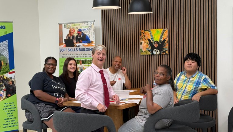 A group of men and women supported in the Project Friendship group meet together around a large wooden table in the Impact Center for Learning. The spirit of friendship shines on their faces.