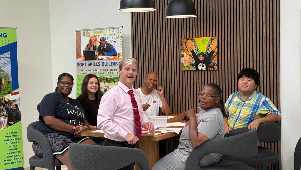 A group of men and women supported in the Project Friendship group meet together around a large wooden table in the Impact Center for Learning. The spirit of friendship shines on their faces.