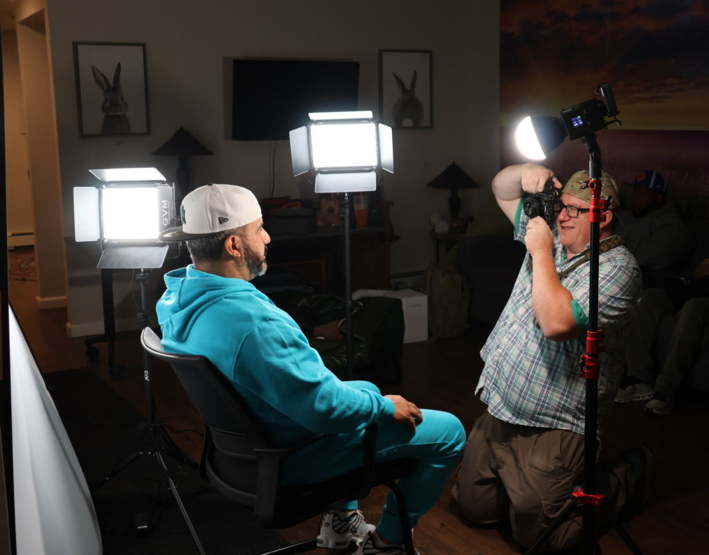 A DSP sits in a chair while having his portrait taken by photographer Jon Harrison. They are surrounded by lighting equipment which glow in the dark room.